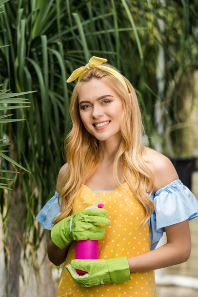 beautiful young woman in rubber gloves holding sprayer and smiling at camera in greenhouse - Foto, imagen