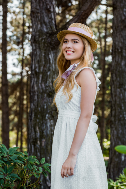 beautiful smiling young woman in dress and wicker hat with ribbon standing and looking away in forest - Fotoğraf, Görsel