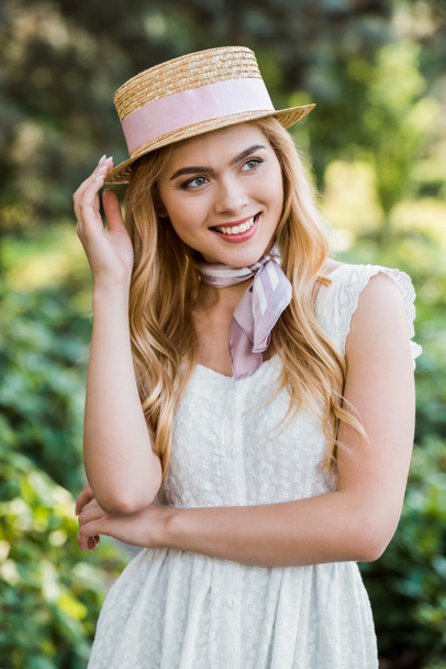 mooi mals blonde meisje in jurk en rieten hoed met lint glimlachen en kijken weg in park  - Foto, afbeelding