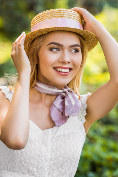 beautiful smiling blonde girl wearing wicker hat with ribbon and looking away in park - Foto, imagen