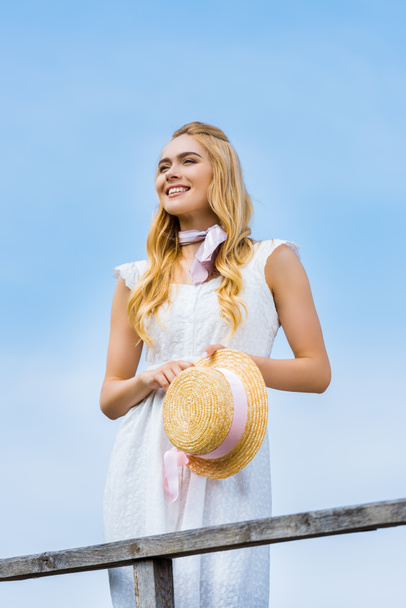 krásná usměvavá blondýnka drží proutěný klobouk s mašlí a hledat dál proti modré obloze - Fotografie, Obrázek