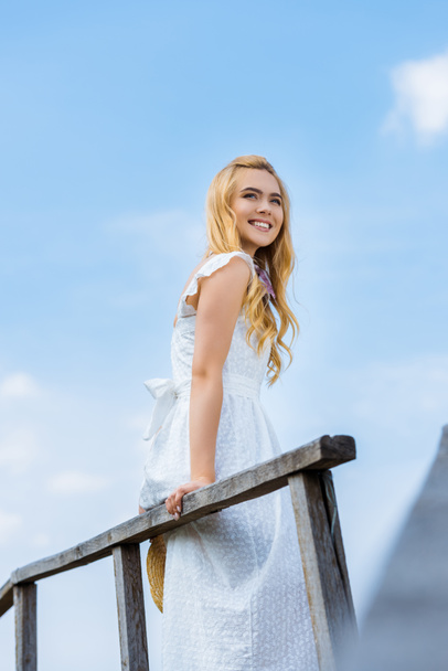 niski kąt widzenia szczęśliwy blond dziewczyna uśmiechając się i patrząc od hotelu przeciw błękitne niebo  - Zdjęcie, obraz