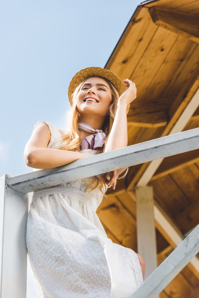 vista de ángulo bajo de la chica feliz en vestido blanco y sombrero de mimbre apoyado en barandilla en el día soleado
 - Foto, imagen