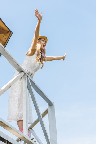 low angle view of cheerful young woman in dress and wicker hat raising hands and looking away against blue sky - Photo, Image
