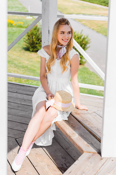 μεγάλη γωνία θέα όμορφη χαμογελαστή κοπέλα με λευκό φόρεμα κάθεται στο ξύλινο πάγκο και αναζητούν μακριά - Φωτογραφία, εικόνα