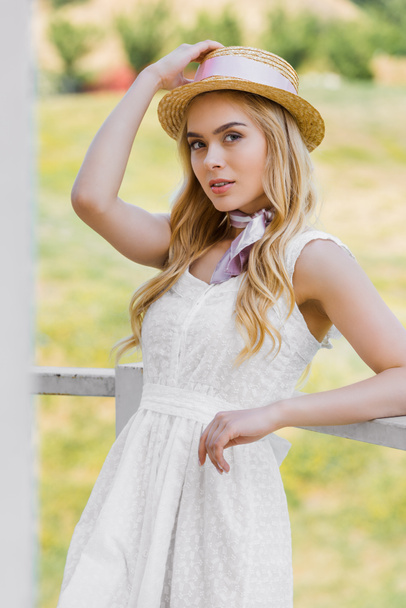 attractive blonde woman in wicker hat and white dress leaning at railing and looking at camera - Foto, imagen