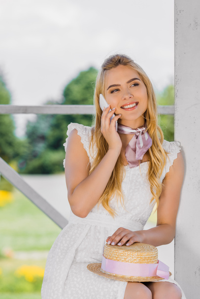 beautiful smiling blonde woman holding wicker hat and talking by smartphone - Fotó, kép