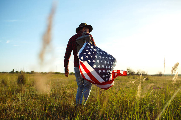 4th of July. American Flag.Patriotic holiday. Traveler with the flag of America. The man is wearing a hat, a backpack, a shirt and jeans. Beautiful sunset light. American style.  - Foto, Imagem