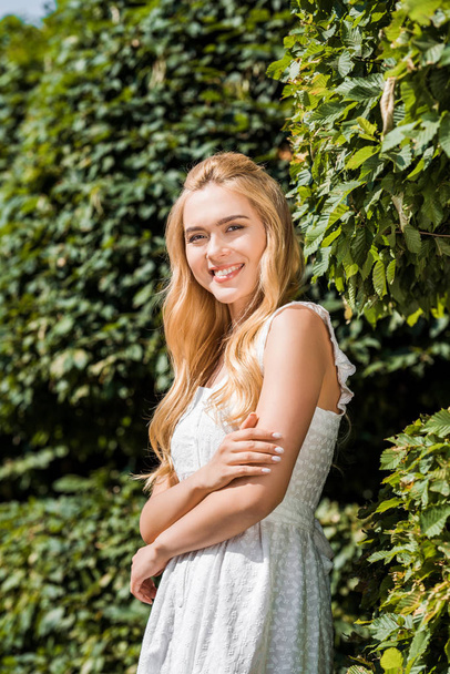 beautiful blonde girl in white dress smiling at camera while standing near green plants - Photo, Image