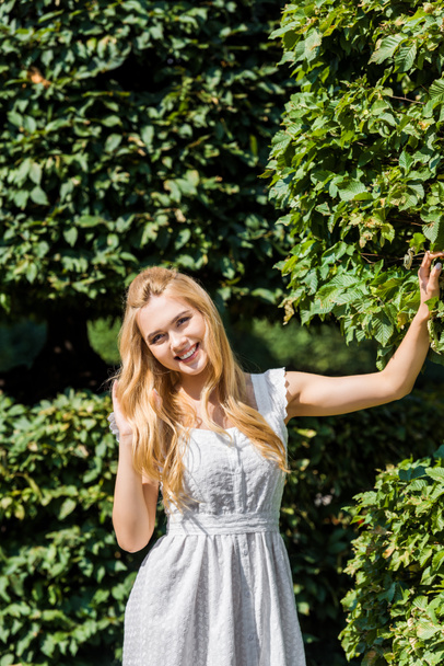 schöne junge Frau im weißen Kleid lächelt in die Kamera, während sie neben grünen Blättern steht - Foto, Bild
