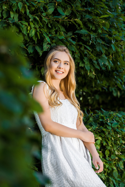 selective focus of beautiful smiling blonde girl in white dress posing near green leaves  - Photo, Image