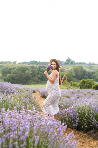 hermosa mujer embarazada sonriente en vestido blanco oliendo flores de lavanda en el campo
 - Foto, Imagen