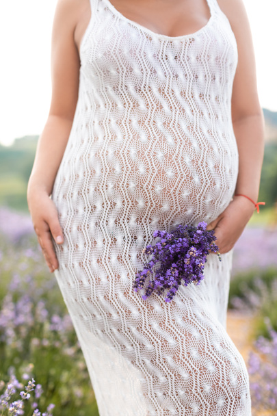 обрезанное изображение беременной женщины, касающейся живота и держащей лавандовые цветы
 - Фото, изображение