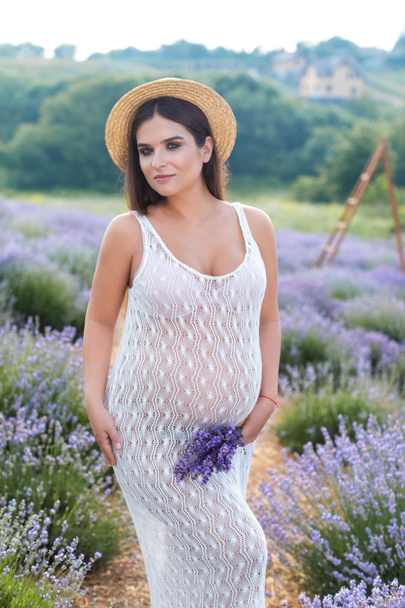 krásná těhotná žena pózuje v bílých šatech na fialové levandule pole a při pohledu na fotoaparát - Fotografie, Obrázek