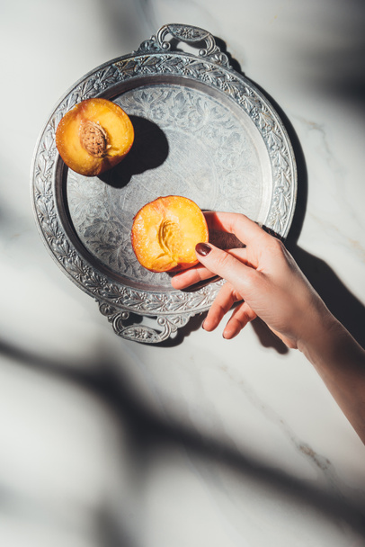 cropped shot of woman holding peach piece on metal tray on light marble surface - Photo, Image