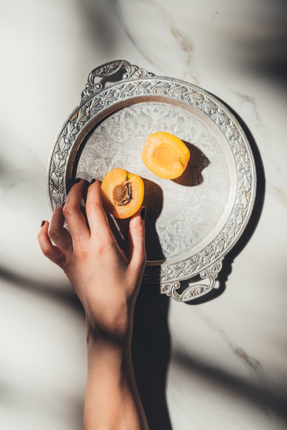 Cropped shot of woman holding apricot piece on metal tray on light marble surface - Photo, Image