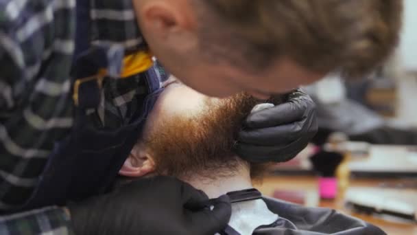 Hairdresser for men. Barbershop. Caring for the beard. A hairdresser with a haircut works over the beard and mustache of a bearded guy. The concept of a hipster lifestyle. Customer Hipster gets a - Кадры, видео