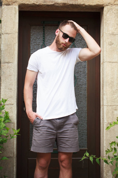 Hipster handsome male model with beard wearing white blank t-shirt and a baseball cap with space for your logo or design in casual urban style - Foto, Imagen