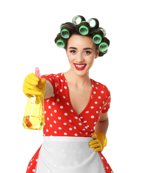 Αστεία νεαρή νοικοκυρά με μαλλιά οδοστρωτήρες κρατώντας απορρυπαντικό σε λευκό φόντο - Φωτογραφία, εικόνα