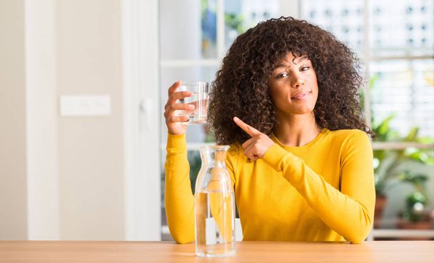 Femme afro-américaine buvant un verre d'eau à la maison très heureux pointant avec la main et le doigt sur le côté
 - Photo, image
