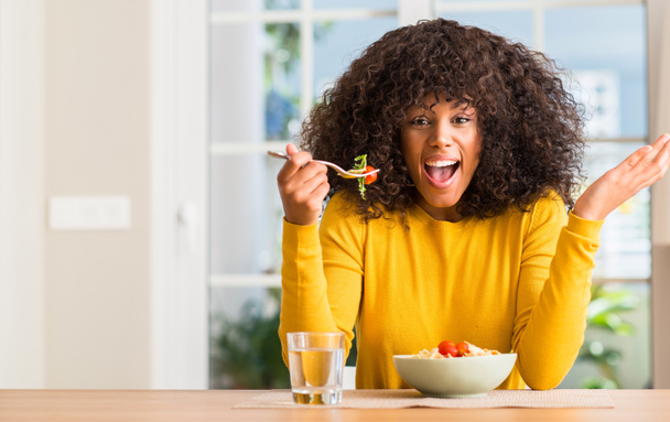 Mujer afroamericana comiendo ensalada de pasta en casa muy feliz y emocionada, expresión ganadora celebrando la victoria gritando con una gran sonrisa y levantando las manos
 - Foto, imagen
