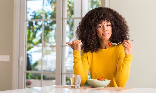 Afrikanische Amerikanerin isst Nudelsalat und zeigt mit der Hand nach oben mit glücklichem Lächeln - Foto, Bild