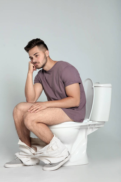 Young man sitting on toilet bowl against gray background - Photo, Image