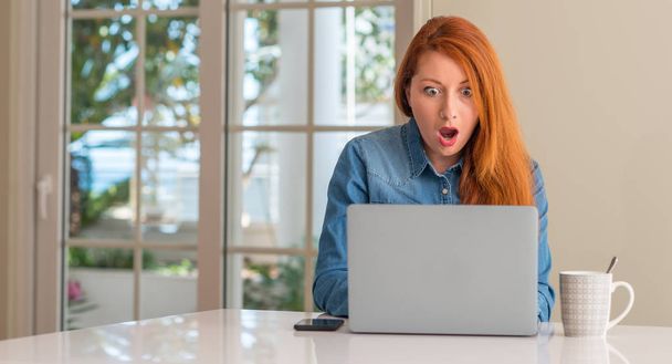 Femme rousse utilisant un ordinateur portable à la maison effrayée sous le choc avec un visage surprise, effrayée et excitée par l'expression de la peur
 - Photo, image