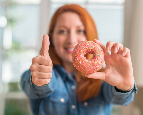 Mulher ruiva segurando donut em casa feliz com grande sorriso fazendo sinal ok, polegar para cima com os dedos, excelente sinal
 - Foto, Imagem