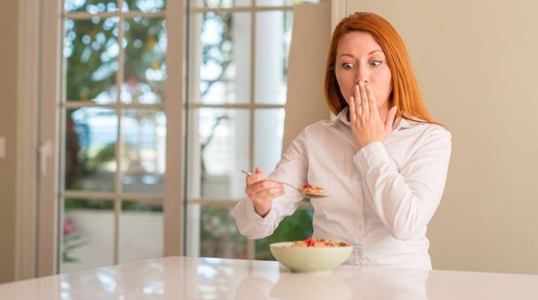 Ruda kobieta jedzenie zbóż z malinami w domu pokrywa usta ręką w szoku ze wstydem za błąd, wyrażenie strachu, boi się w milczeniu, tajne koncepcja - Zdjęcie, obraz