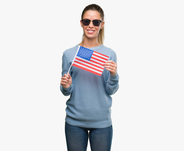 ABD bayrağı ile ayakta ve diş gösterilen kendine güvenen bir gülümsemeyle gülümseyerek mutlu bir yüz tutan güzel genç kadın - Fotoğraf, Görsel