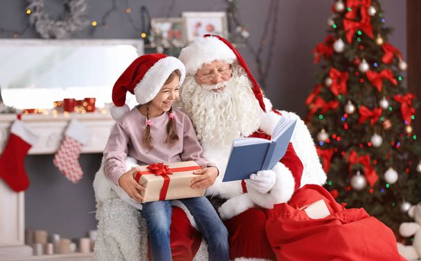 Authentic Santa Claus reading book to little girl indoors - Foto, Bild