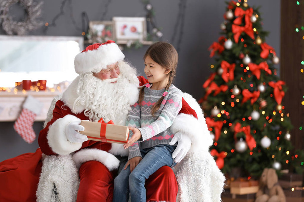 Little girl with gift box sitting on authentic Santa Claus' lap indoors - Fotografie, Obrázek