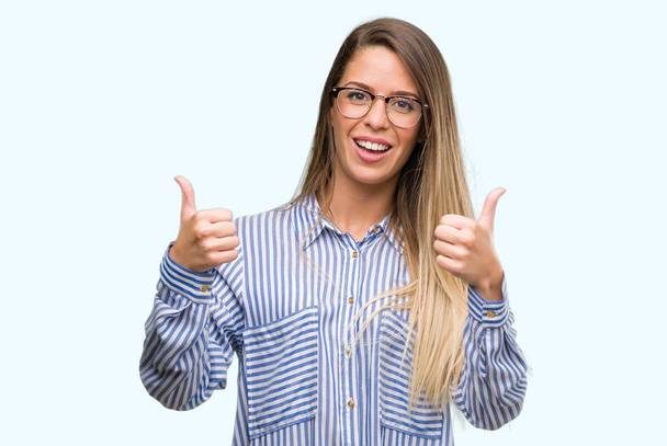 Όμορφη νεαρή γυναίκα που φοράει κομψό πουκάμισο και τα γυαλιά σημάδι επιτυχίας κάνει θετική κίνηση με το χέρι, thumbs up χαμογελαστός και χαρούμενος. Κοιτάζοντας την κάμερα με χαρούμενη έκφραση, χειρονομία νικητής. - Φωτογραφία, εικόνα