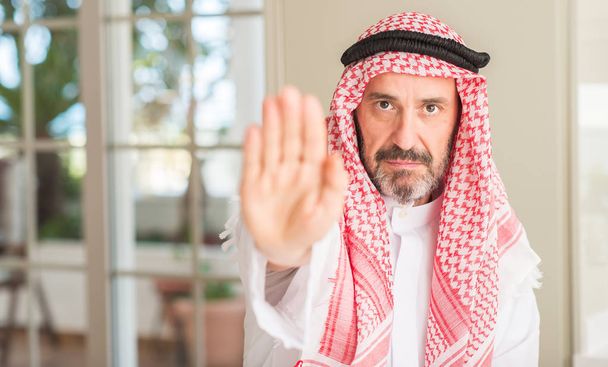 Homem árabe de meia idade em casa com a mão aberta fazendo sinal de parada com expressão séria e confiante, gesto de defesa
 - Foto, Imagem
