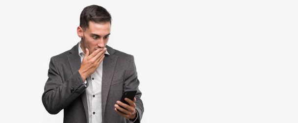 Knappe jongeman met smartphone cover mond met hand geschokt met schaamte voor fout, uitdrukking van angst, bang in stilte, geheime concept - Foto, afbeelding