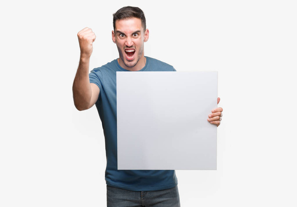 Handsome young man holding advertising banner annoyed and frustrated shouting with anger, crazy and yelling with raised hand, anger concept - Photo, image