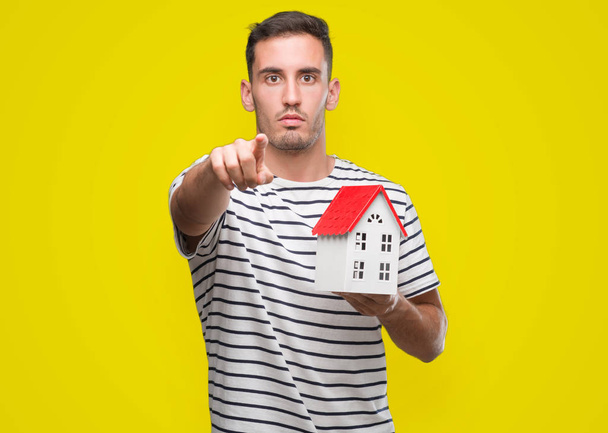 Bonito agente imobiliário segurando uma casa apontando com o dedo para a câmera e para você, sinal de mão, gesto positivo e confiante da frente
 - Foto, Imagem