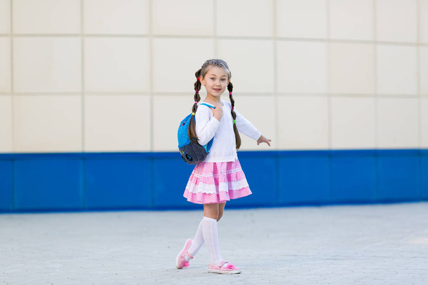 A little beautiful girl in a summer dress with a backpack is standing by the wall in the city. The girl is a young fashionista posing. - Zdjęcie, obraz