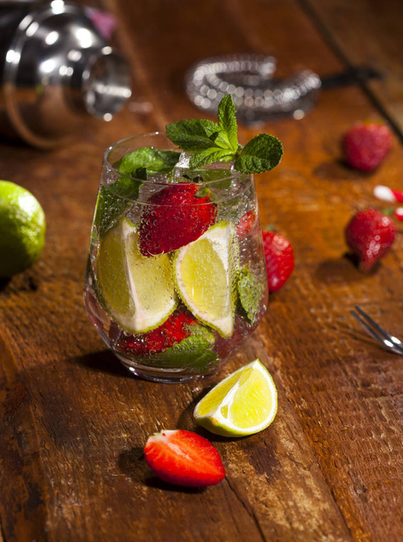 Summer cold drink with strawberries, mint, lime and ice in glass on wooden bar counter. Closeup of cocktail with fresh berry fruits. - Photo, image