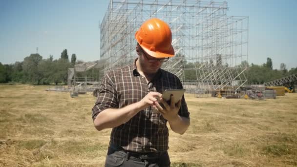 Young builder in helmet standing with tablet on straw field, metal installation on background, concentrated - Imágenes, Vídeo