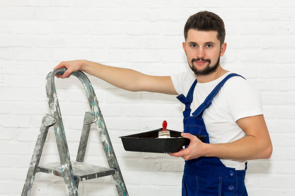 Young man with dark hair is painting the wall in white in his apartment. In his hand he holds a paint pan and paint brush, leaning against the ladder. He looks at the viewer and smiles. - 写真・画像