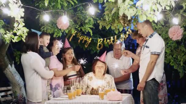 Grote familie vieren verjaardag thuis in de tuin in de avond. Vrouw kaarsen op een taart uitblazen - Video