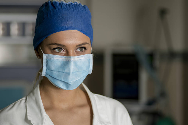 Joven enfermera rubia en sala de emergencias con máscara quirúrgica y gorra quirúrgica
 - Foto, Imagen