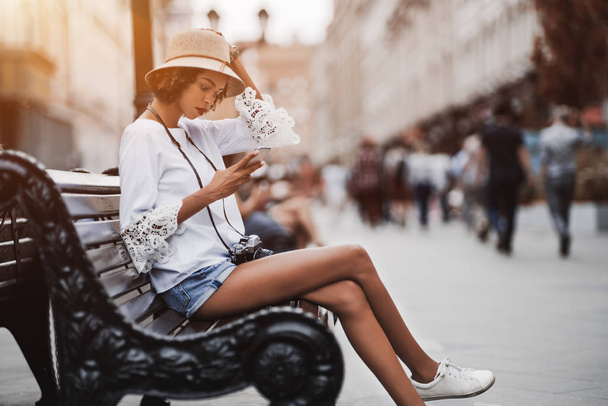 Pamlsek afroamerické žena v bílé košili a klobouk a s fotoaparátem retro film sedí venku na dřevěné lavici rušné večerní ulice a s konverzaci s rodinou přes smartphone - Fotografie, Obrázek