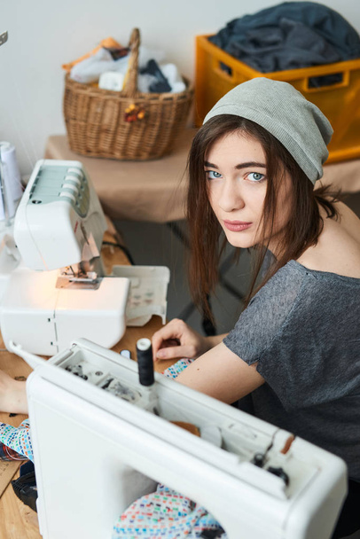 attractive brunette dressmaker in hat sewing cloth with sewing machine in atelier - Foto, Imagem