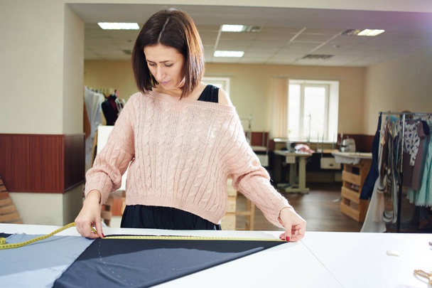 brunette seamstress with measuring tape measuring textile on table in atelier - Fotografie, Obrázek