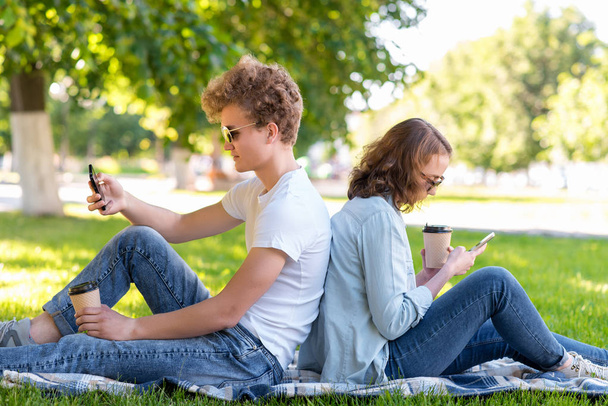 A young couple. Summer in nature. A boy with a girl resting on a plaid. Holds hot coffee or tea. They look at smartphones. Write messages on social networks. - Foto, imagen