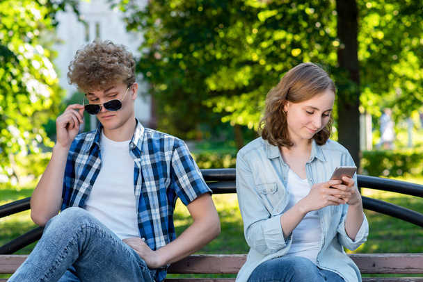 The guy and the girl are sitting on bench. Summer in nature. A girl in her hands holds a smartphone communicates in social networks. The guy spies Through glasses behind the girl. - Фото, изображение