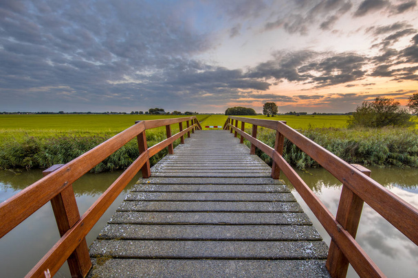 Puente de madera sobre el canal en el paisaje agrícola contra la hermosa puesta de sol de colores
 - Foto, imagen